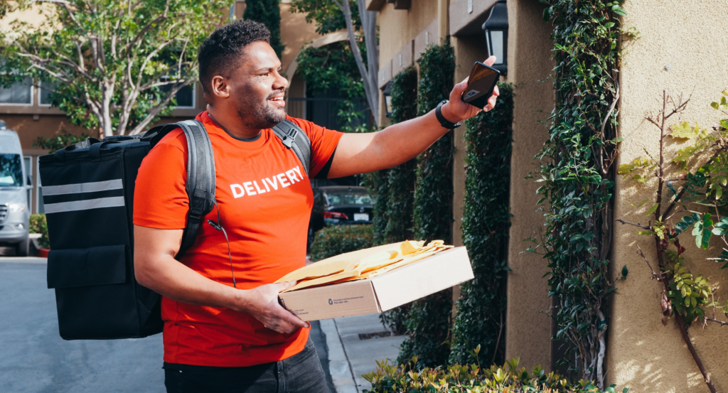 A delivery person has a black insulated backpack on their back and is smiling as they use their phone