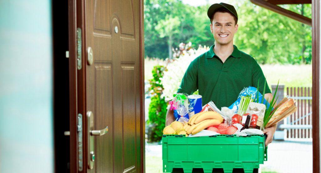 A cheerful delivery person in a green uniform stands at an open doorway, holding a green crate filled with various groceries, including bananas, bread, eggs, vegetables, and other packaged items.